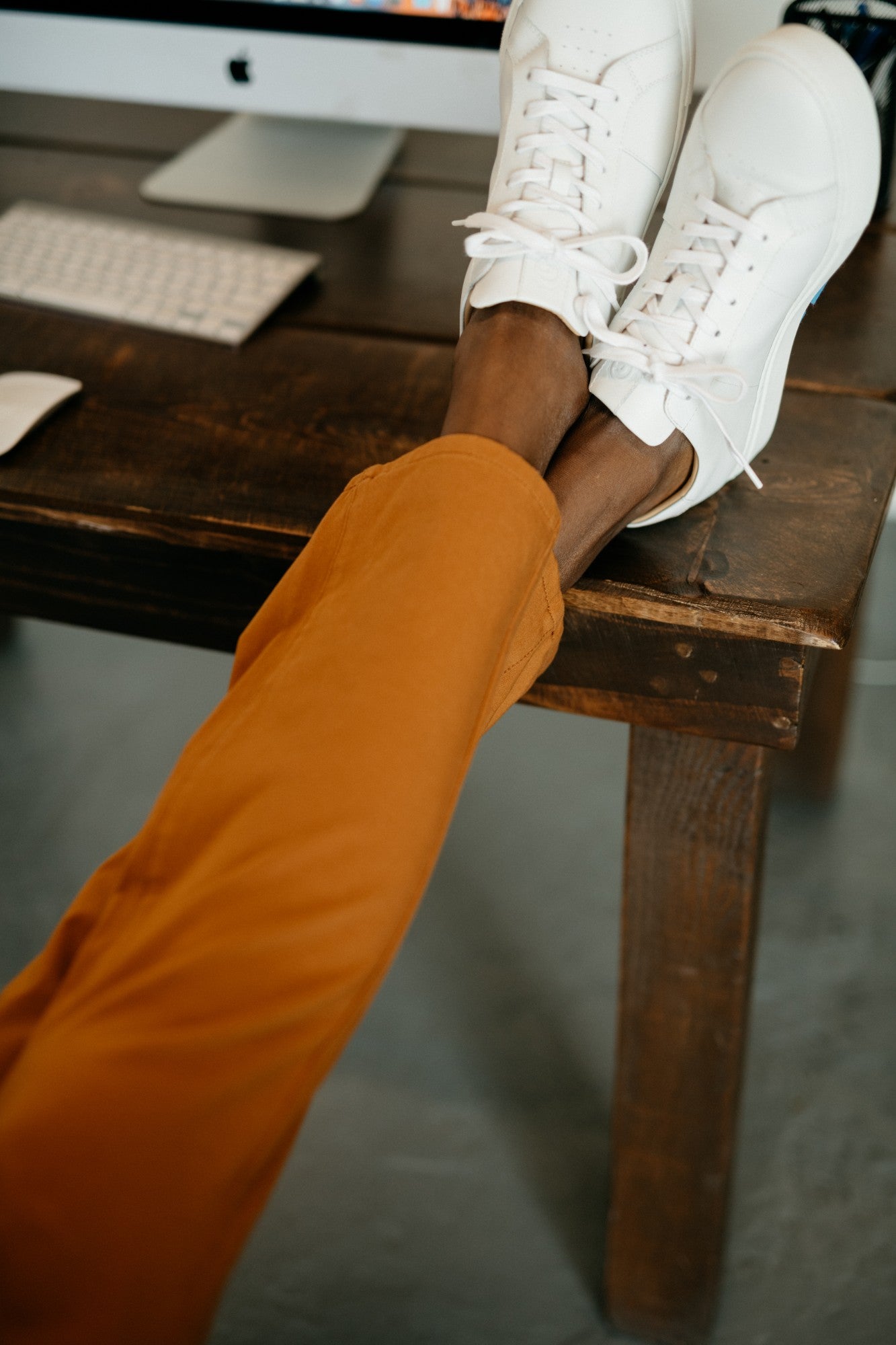 Person wearing white sneakers and orange pants sitting on a wooden desk.